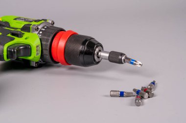 Bir matkap gri bir yüzeyde duruyor ve yanında da tornavida parçaları var. Matkabın kırmızı yakalı siyah ve yeşil bir tasarımı var. Bitler matkap tarafından küçük bir grupta düzenlenir..