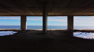 Sahne, beton sütunların çerçevelediği sakin okyanus manzarasını büyük bir çıkıntının altında yakalıyor. Yerdeki su birikintileri, kıyı boyunca sessiz bir an sunan parlak gökyüzünü yansıtır..