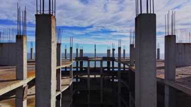 Havadan bakıldığında, okyanusa bakan eksik bir yapı görülüyor. Beton sütunlar mavi gökyüzü ve bulutlardan oluşan bir zemin üzerinde yükseliyor, inşaat ilerlemesini vurguluyor.