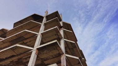 Açık mavi gökyüzüne karşı yükselen beton bir binanın bulunduğu inşaat alanı. Açığa çıkmış kirişler ve tamamlanmamış bölümler devam eden inşaat çalışmalarını gösteriyor.