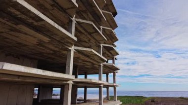İhmal edilmiş beton bir yapı denizin kenarında duruyor, sakin bir arkaplan ve unutulmuş bir geçmişe işaret eden geniş, mavi bir gökyüzüne karşı çıplak iskeletini sergiliyor..