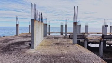 Bu hava perspektifi, deniz kenarında terk edilmiş bir inşaat alanını gösteriyor. Beton sütunlar dik dururken okyanus bulutlu bir gökyüzünün altında uzanıyor..