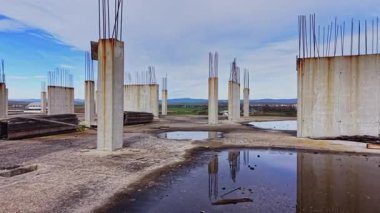Su birikintileri bitmemiş bir inşaat alanında gökyüzünü yansıtıyor. Kırsal bölgelerdeki gelişmeyi durduran, çorak bir arazinin ortasında uzun sütunlar durmaktadır..