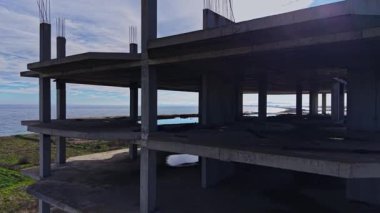 Sahne, okyanusa bakan ve destek kirişleri görünen bitmemiş beton bir bina gösteriyor. Yeşil bitki örtüsü, açık bir gökyüzünün altındaki yapıyı sarar..