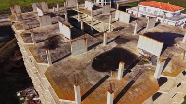 İnşaat alanının hava görüntüsü, inşaat demiri ve paslanmış kirişleri olan eksik bir bina gösteriyor. Çevreleyen alanlar ve bir yol olay yerine bağlam sağlıyor.