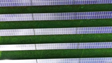 Güneş panellerinin hava görüntüsü düzgün sıralanmış, canlı yeşil bir alana karşı duruyor. Bu tesis, parlak gün ışığında yenilenebilir enerjiyi kullanır..