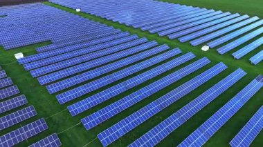 Geniş bir güneş paneli alanı yeşil bir arazide güneş ışığını yakalar. Mavi paneller genişliyor, yenilenebilir enerji faaliyetlerini sergiliyor.