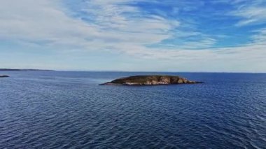 Görüntü, uçsuz bucaksız mavi okyanusun ortasındaki küçük yeşil bir adayı gösteriyor. Gökyüzü parçalı bulutlu, bu uzak yer için sakin bir atmosfer yaratıyor..