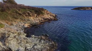 Kayalık kayalıklar bu hava manzarasında sakin mavi denizle buluşur. Kıyı şeridinde çeşitli doku ve renkler yer alıyor ve sahil kenarında huzurlu bir deniz manzarası yaratıyor..