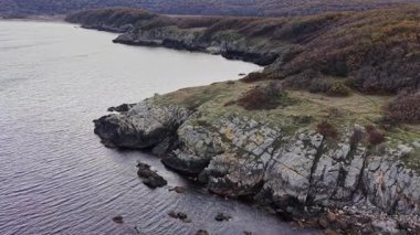 Kayalık kayalıkların ve renkli sonbahar ağaçlarının bulunduğu engebeli bir kıyı şeridinin havadan görünüşü bulutlu bir gökyüzünün altında yavaşça durgun sulara doğru eğimli..