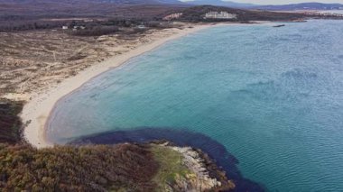 Bu hava manzarası sakin kumlu bir kumsalın sakin mavi sularla buluşmasını yakalıyor. Yuvarlanan tepeler arka planda görünür, barışçıl bir kıyı manzarası yaratır..