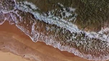 Dalgalar sıcak kumlu bir plaja yuvarlanarak parlak güneş ışığı altında berrak mavi sularla güzel bir kontrast oluşturuyor. Kıyı şeridi davetkar bir şekilde kıvrılır.