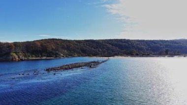 Bu hava görüntüsü, kayalık oluşumların ve açık mavi gökyüzünün altındaki yemyeşil tepelerin yer aldığı sakin bir kıyı şeridini sergiliyor. Doğanın güzelliğini yansıtıyor..