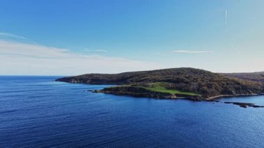 Parlak mavi sular kıyı şeridine kadar uzanıyor ve berrak bir gökyüzünün altındaki yemyeşil tepeleri ortaya çıkarıyor. Bu sakin sahil manzarası doğanın güzelliğini gündüz saatlerinde yakalar..
