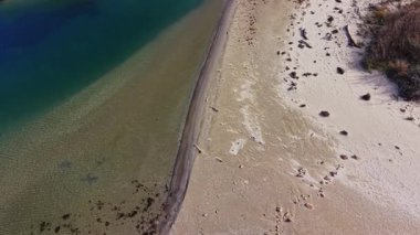 Bu hava manzarası, yumuşak kumların sakin ve berrak sularla buluştuğu sakin bir sahil şeridini yakalar ve parlak gün ışığında huzurlu bir doğal manzarayı gözler önüne serer..