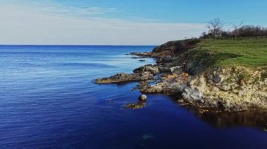 Uçurumların üzerindeki yemyeşil çimlerle mavi sularla buluşan kayalık bir kıyı şeridinin havadan görünüşü. Sahne sakin ve el değmemiş, doğa aşıkları için mükemmel..
