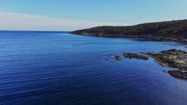 Bu hava manzarası engebeli bir kayalık sahil şeridi boyunca mavi denizin sükunetini yakalar. Açık gökyüzü ufuk boyunca uzanıyor, huzurlu atmosferi güçlendiriyor..