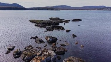 Hava manzarası bulutlu bir gökyüzünün altında hareketsiz sulara uzanan kayalık bir kıyı şeridini yakalar. Huzurlu bir günde arka planda kayan tepeler görünür..
