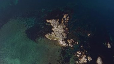 Hava manzarası canlı mavi sularla çevrili kayalık bir kıyı şeridini yakalar. Güneş ışığı yüzeyden yansıyarak bu kıyı bölgesinin doğal güzelliğini vurgular..