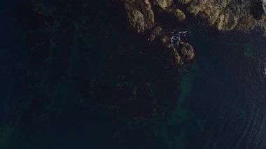 Yukarıdan, engebeli kıyı şeridi derin mavi okyanusla buluşuyor, kayalık oluşumlar ve berrak sular sergiliyor, bu sakin manzaradaki doğanın güzelliğini vurguluyor..