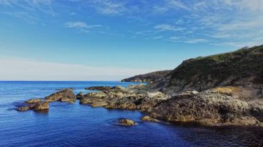 Bu hava perspektifi, berrak suları olan güzel kayalık bir sahil şeridini gösteriyor. Yumuşak dalgalar, güneşli, mavi bir gökyüzünün altında kıyıya doğru süzülür ve dingin bir manzara yaratır..