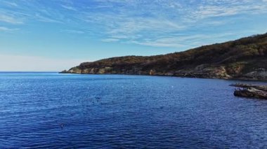 Bu hava sahnesi, yumuşak dalgaların kıyıya vurduğu sakin bir kıyı şeridi yakalıyor. Bereketli yeşil tepeler ve berrak mavi gökyüzü huzurlu bir atmosfer yaratır..
