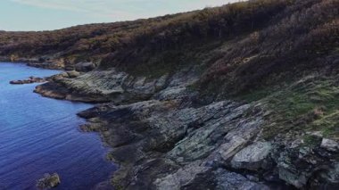 Hava perspektifi kayalar ve seyrek bitkilerle kaplı engebeli bir kıyı şeridini yakalar. Sakin sular, berrak bir gökyüzünün altındaki kayalık araziyle tezat oluşturuyor..