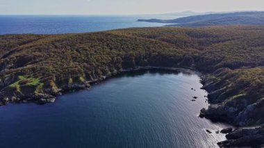 Tepeden çekimde yoğun yeşil ağaçlar ve açık bir gökyüzünün altında kayalık kıyılarla çevrili sakin bir koy görülüyor. Kıyı şeridinin doğal güzelliğini vurguluyorlar..