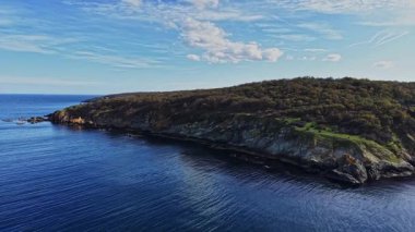 Bu hava manzarası, yemyeşil, kayalık uçurumlar ve yukarıdaki berrak gökyüzünü yansıtan sakin bir kıyı manzarası sergiliyor..