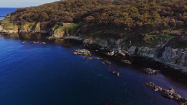 Sersemletici hava manzarası yeşil tepelerle çevrili berrak mavi sularla kayalık bir kıyı şeridini gösteriyor. Kıyı şeridi benzersiz kaya oluşumları ve sakin manzaraya sahiptir..