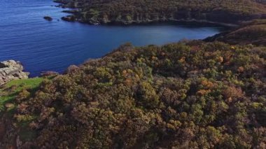 Sonbahar yeşilliklerinin canlı renkleri yukarıdan çekilen barışçıl bir gölü çevrelemektedir. Yumuşak dalgalar sakin bir manzarada kayalık kıyı şeridine çarpar..