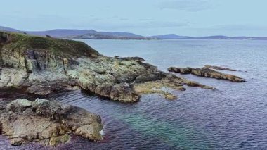 Bu hava perspektifi kayalık oluşumları, sakin suları ve berrak bir gökyüzü altındaki uzak tepeleriyle engebeli bir kıyı şeridini sergiliyor, manzaranın huzurlu güzelliğini yansıtıyor..