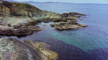 Bu hava görüntüsü kayalık oluşumları ve berrak mavi sularıyla engebeli bir kıyı şeridini yakalıyor. Huzurlu manzara, parlak bir gökyüzünün altında doğal güzellikleri sergiliyor..