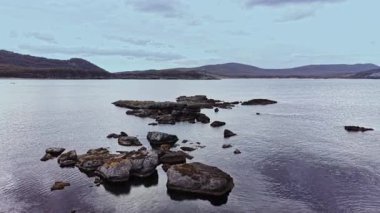 Gün batımında bulutlu bir gökyüzünün altında sakin sularla çevrili kayalıklarla ve uzak dağlarla çevrili kayalık bir kıyı şeridini görüntüler..