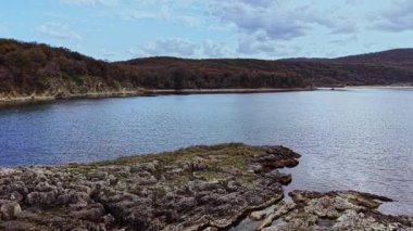 Bu hava çekimi, engebeli kayalar ve tepelerle çevrili huzurlu bir su kütlesi yakalar ve parçalı bulutlu gökyüzünün altında doğanın güzelliğini gösterir..