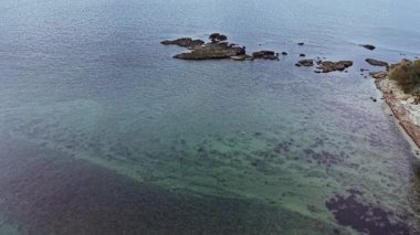 Hava görüntüsü, kıyı boyunca uzanan kıyı bitki örtüsüyle altındaki kayaları ortaya çıkaran berrak suları gösteriyor. Durgun su, manzaranın doğal güzelliğini arttırır..