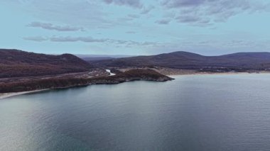 Bu hava sahnesi, doğanın renklerini yansıtan sakin suları barındıran bulutlu bir gökyüzünün altında yeşil tepelerle çevrili nazik dalgalarla dolu güzel bir kıyı şeridini sergiliyor..