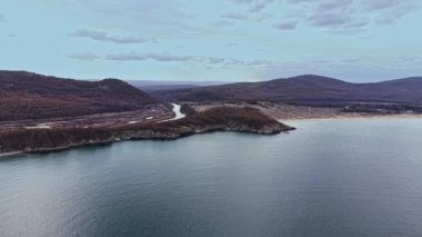 Tepelerle çevrili huzurlu bir sahil şeridinin güzelliğini gösteren bir hava manzarası. Sakin sular gökyüzünü yansıtarak, bölgenin gündüz boyunca doğal ihtişamını vurguluyor..