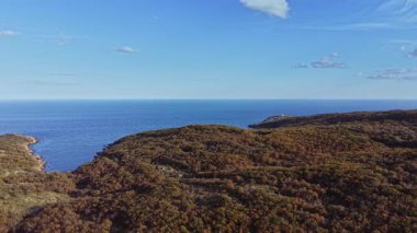 Geniş kıyı arazisi, yemyeşil tepelerin berrak bir gökyüzünün altında sakin mavi denizle buluştuğunu gösteriyor. Bu sahne gün ışığında doğanın güzelliğini yakalar..