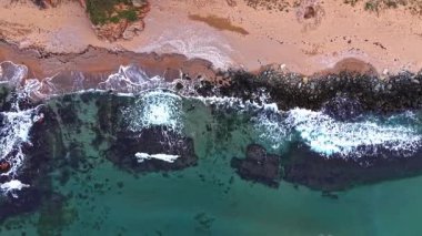 Bu hava manzarası, turkuaz suyu, kayalık kıyı şeridini ve plajda köpük oluşturan yumuşak dalgaları vurgulayan güzel kıyı şeridini sergiliyor, sakin bir atmosferi yakalıyor..
