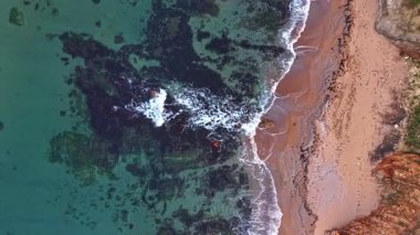 Kayalara vuran dalgalarla kumlu bir plaj manzarası. Açık mavi su kıyıya vurur, deniz tabanının ve kıyı manzarasının dokusunu ortaya çıkarır..