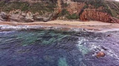 Kayalıklar yükselirken dalgalar yavaşça kıyıya çarpar. Sahil sakin görünüyor, turkuaz berrak su kumlu toprakla buluşuyor..