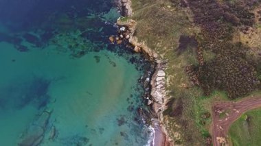 Havadan bakıldığında engebeli uçurumların turkuaz sularla buluştuğu sakin bir sahil sahnesinde kumlu sahil boyunca dağılmış çakıl taşları görülür..