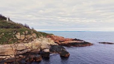 Aerial view features a rugged coastal landscape with colorful rocks and calm waters. Soft clouds hover above, creating a peaceful atmosphere near the shore.