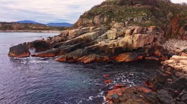 Hava perspektifi engebeli bir kıyı şeridini yakalar ve büyük kayalar durgun sulara saplanır. Çevresindeki tepeler bulutlu bir gökyüzünün altında manzaralı bir zemin sağlar..