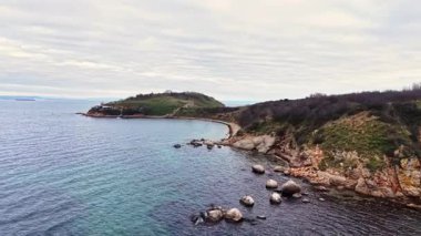 Bir hava manzarası kayalık kıyı şeridini, yeşil tepeleri ve sakin mavi suları yakalar. Sahne huzurlu, bulutlu bir günbatımında doğanın güzelliğini gösteriyor..