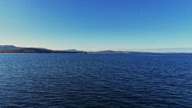 Uçsuz bucaksız mavi deniz, ufuk boyunca görünen uzak tepelerle berrak bir gökyüzünün altında uzanır. Sakin su, manzaranın sakin atmosferini yansıtır..
