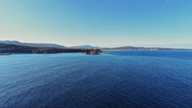 Geniş mavi sular, berrak bir gökyüzünün altındaki uzak tepelere doğru uzanıyor. Huzurlu manzara, doğada gün boyunca sakin bir anı yakalar..