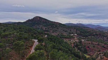 Manzara İspanya 'daki bir dağ manzarasını gösteriyor. Dolambaçlı bir yol bulutlu bir gökyüzünün altındaki yeşil ağaçların ve tepelerin arasından geçer. Sahne doğayı ve yolu yukarıdan yakalıyor..