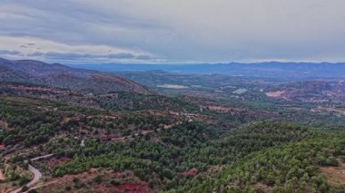 Geniş bir manzara İspanya 'nın kayan tepelerini ve vadilerini gösteriyor. Çevrede yeşil ağaçlar ve kayalık alanlar var. Gökyüzü bulutlu, yaklaşan hava değişimine işaret ediyor..